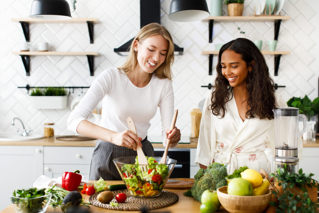 two women different nationalities are smiling cooking salad kitchen